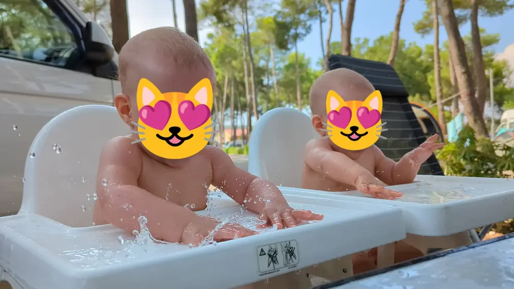 Our two baby daughters outside in high chairs with tables attached. They're playing with water we've spilled on the tables. You see water squirting in alle directions where they're hitt
ing the table. Faces hidden with cat with heart eye emojis.