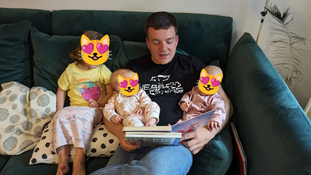 Me sitting on a sofa reading a book with my 3 year old daughter to the right and two babies on my lap. Their faces hidden with emojis.